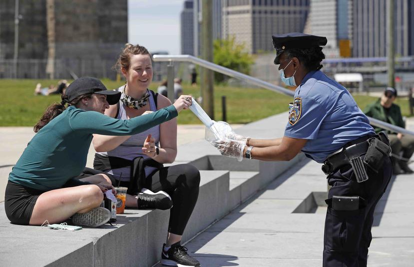 Una oficial de seguridad escolar del Departamento de Policía de Nueva York entrega máscaras faciales a dos mujeres en el Brooklyn Bridge Park, en Nueva York. (AP)