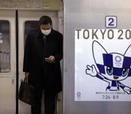 Un cartel de los Juegos Olímpicos de Tokio 2020 es visto junto a la puerta un tren del metro mientras un viajero con una máscara mira su celular. (AP)