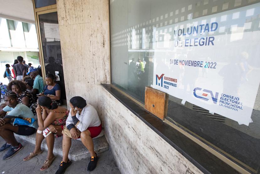 Varias personas permanecen sentadas junto a un cartel que anuncia las elecciones locales del 27 de noviembre, en La Habana, Cuba.