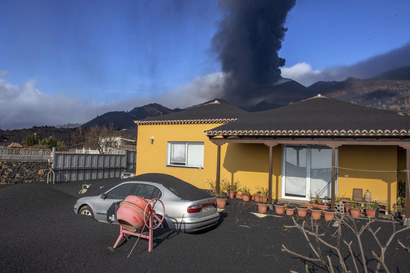Una capa de ceniza volcánica cubre una casa, un automóvil y sus alrededores mientras en el fondo un volcán sigue en erupción en la isla canaria de La Palma, España.