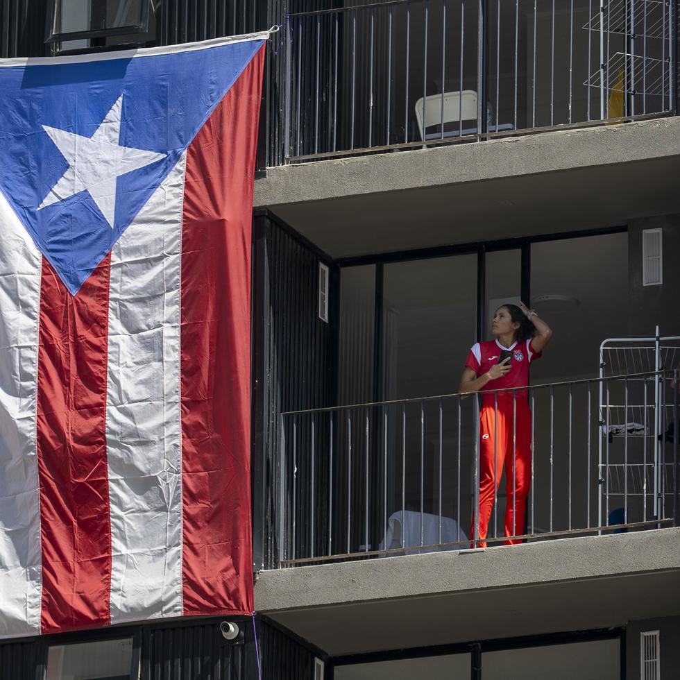 La fondista Beverly Ramos es apreciada en el balcón de su habitación de la Villa Panamericana, mientras observa la enorme bandera puertorriqueña que cuelga de los balcones.