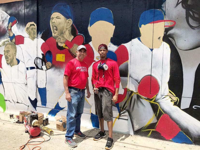 El alcalde de Vega Alta, Oscar Santiago, junto al artista Abdiel Vázquez, posan frente al mural en la calle principal del centro del pueblo.