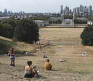 Gente sentada en pasto seco por el sol en el Parque de Greenwich, con el Museo Marítimo y el distrito financiero de Canary Wharf al fondo, en Londres.