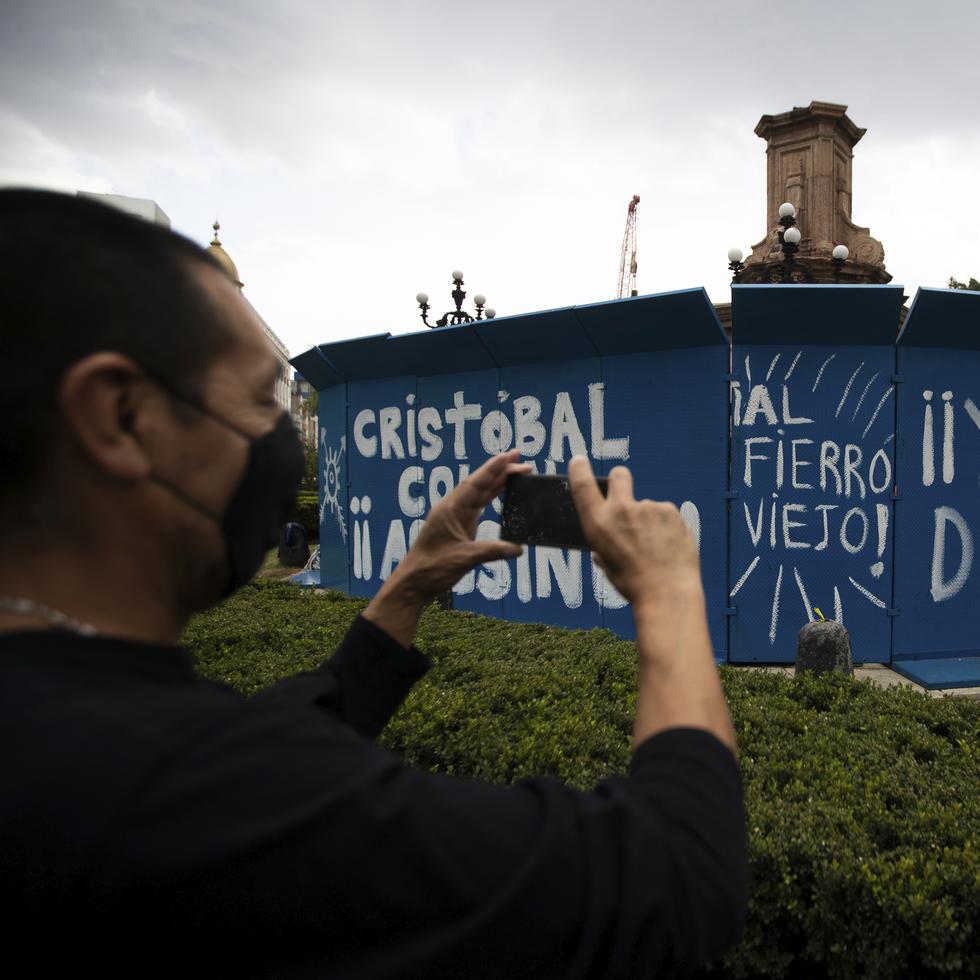 Un peatón toma una foto de unas pintadas en rechazo a Cristóbal Colón realizadas sobre unas barreras de metal que protegen el perímetro del lugar en el que estaba la estatua, en 2020.