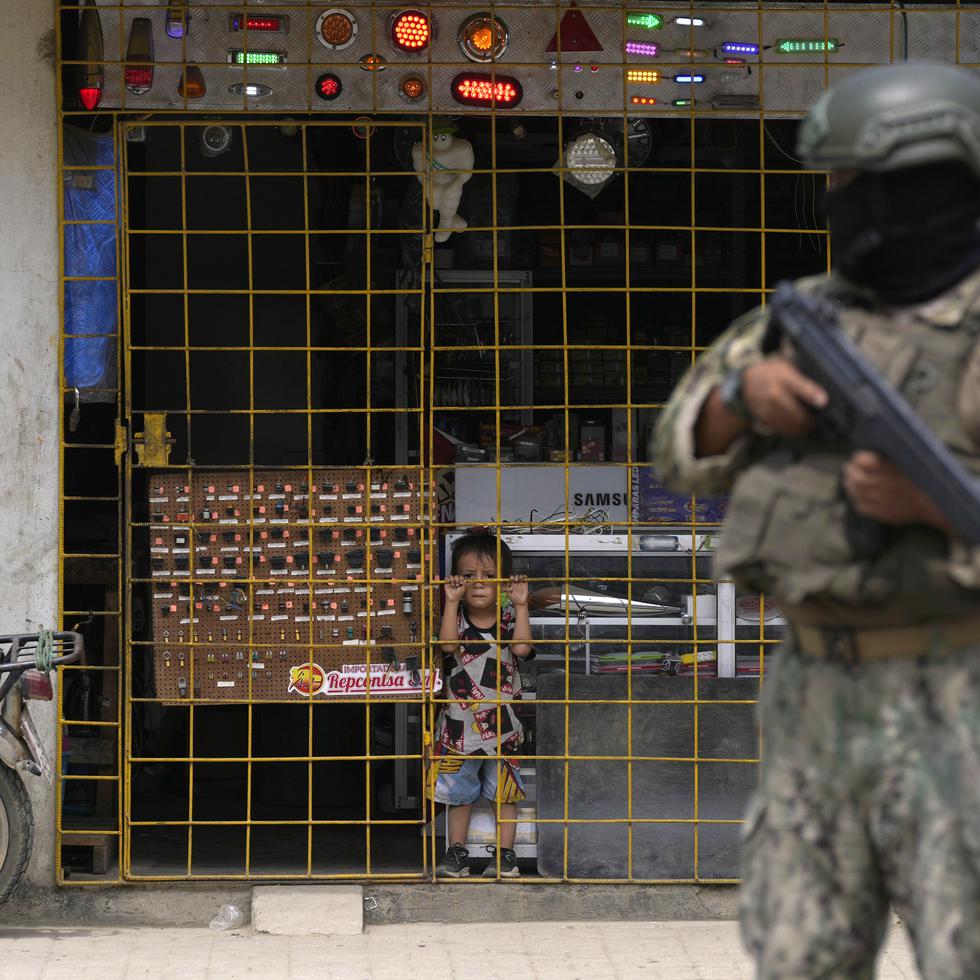 A boy watches from behind the bars of an open electronics store as a soldier stands guard at a security check point placed by the army in Duran, across a bridge from Guayaquil, Ecuador, Monday, Aug. 14, 2023. Ecuador's president declared a state of emergency in some areas after a presidential candidate was killed at a rally ahead of snap elections, set for Aug. 20, after President Guillermo Lasso dissolved the National Assembly to avoid being impeached. (AP Photo/Martin Mejia)