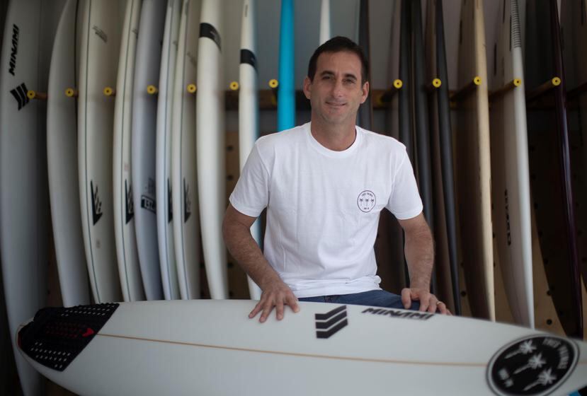 El propietario de Tres Palmas, Carlos Cabrero, posa con la tabla que surfea todas las mañanas antes de ayudar a otros a escoger la suya.