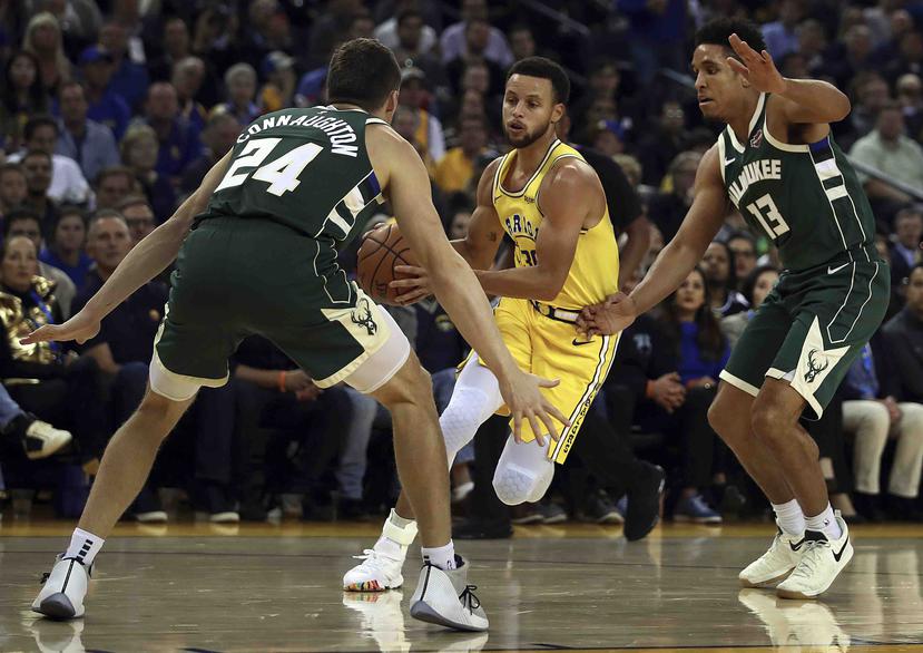 El jugador de los Warriors de Golden State Stephen Curry (centro) conduce el balón entre Pat Connaughton (24) y Malcolm Brogdon, de los Bucks de Milwaukee, durante la primera parte del juego de la NBA que enfrentó a ambos equipos, el 8 de noviembre de 201