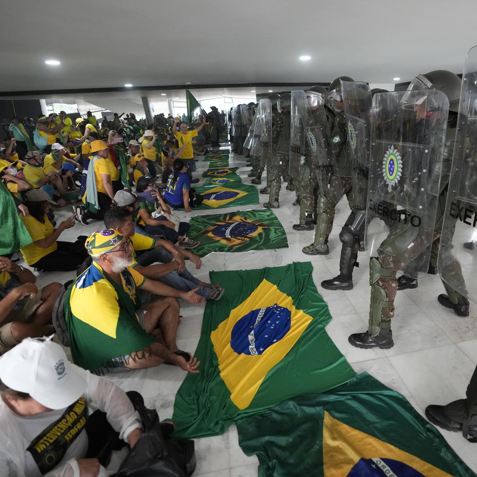 Simpatizantes del expresidente brasileño Jair Bolsonaro permanecen sentados frente a una hilera de policías militares dentro del Palacio de Planalto, luego de irrumpir al interior el domingo 8 de enero de 2023, en Brasilia, Brasil.