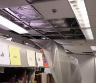 La foto muestra estantes con libros cuya única protección del agua y sucio generado por el sistema de aire acondicionado en el techo expuesto son pedazos de tela de plástico.