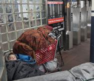 Una persona sin hogar duerme debajo de mantas junto a un carrito de compras lleno de pertenencias en una estación del metro de Nueva York.