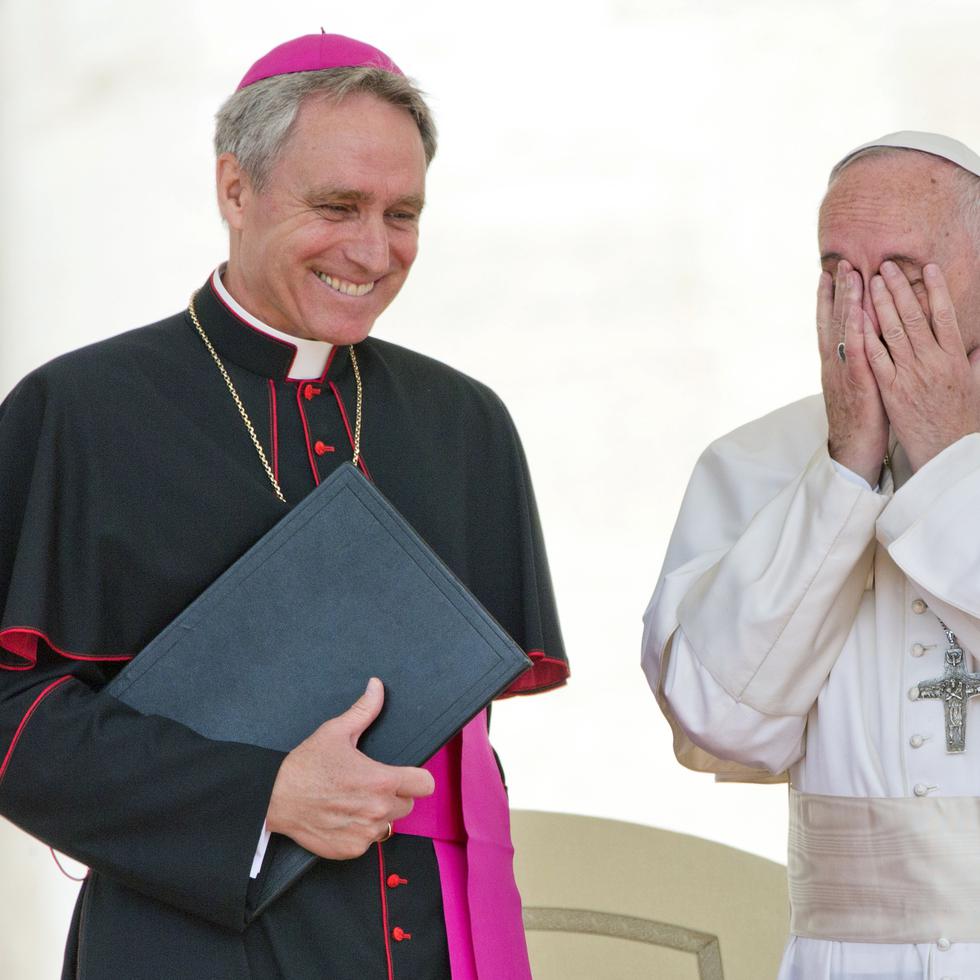 El papa Francisco se lleva las manos a la cara mientras el secretario personal del fallecido papa emérito Benedicto XVI sonríe.