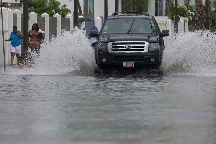Una guagua pasa por un área inundada, mientras unas mujeres se mojan mientras caminan por la acera. (GFR Media)