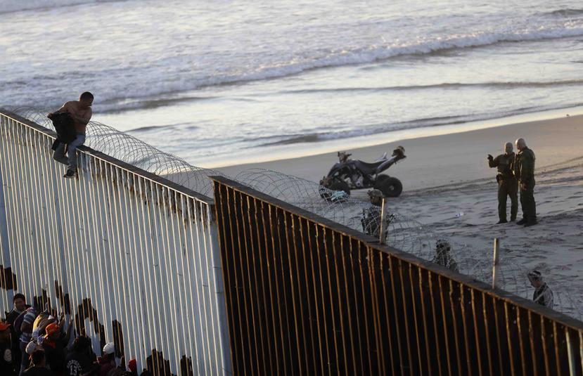 Un migrante centroamericano está sentado sobre la estructura fronteriza que separa a México (izquierda) de Estados Unidos (derecha), mientras agentes fronterizos estadounidenses vigilan la escena. (AP / Gregory Bull)