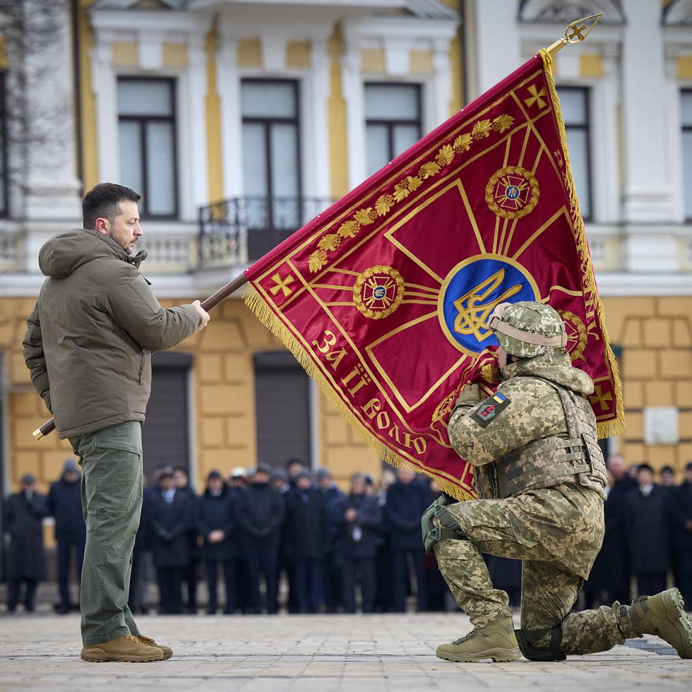 Volodymyr Zelensky, izquierda, sostiene una bandera de una unidad militar ucraniana, mientras un soldado la besa como parte de la ceremonia que conmemora el primer aniversario de la invasión rusa.