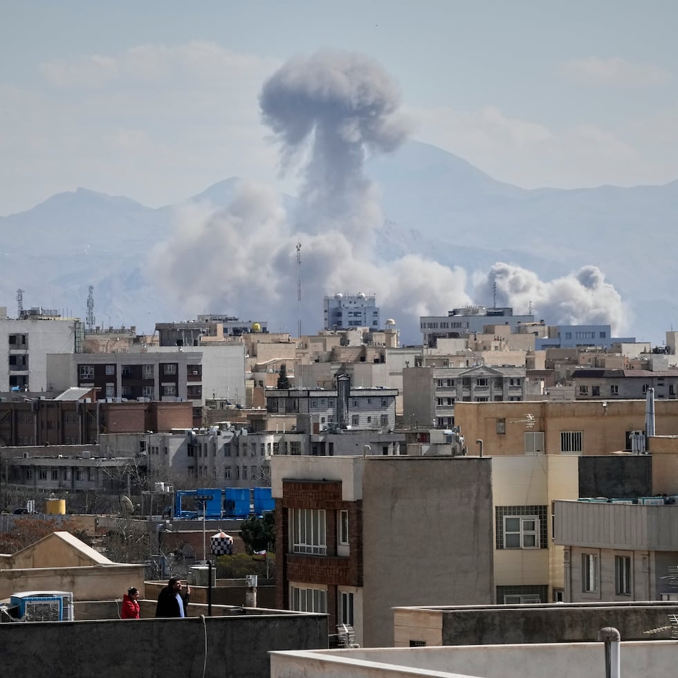 Personas observan desde una azotea mientras una columna de humo se eleva tras un ataque en Teherán, Irán, el domingo 1 de marzo de 2026.