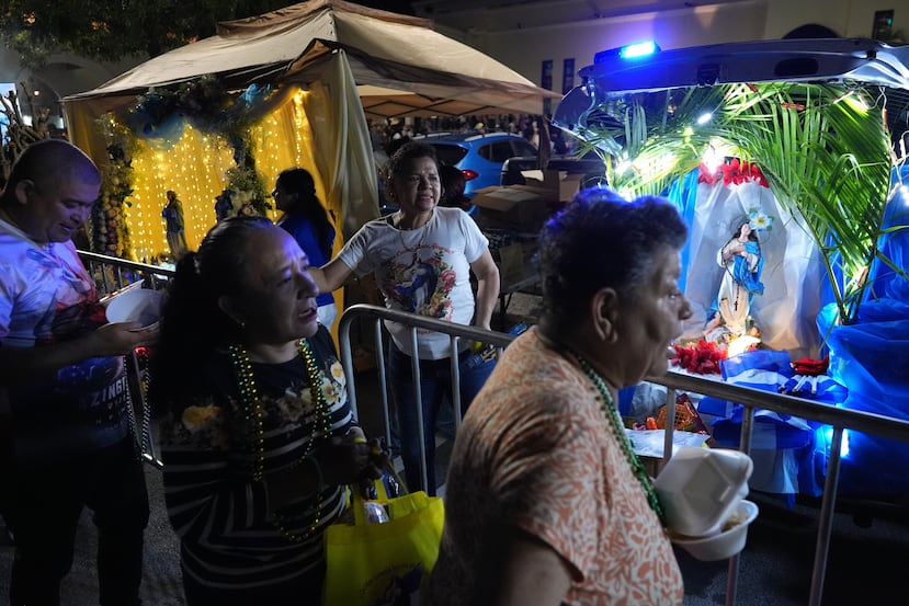 Scarlet Desbas, en el centro, reparte regalos mientras los visitantes cantan en el altar de su familia dedicado a la Virgen María, frente a la Iglesia Católica de San Juan Bosco, durante las celebraciones de la Inmaculada Concepción, el domingo 7 de diciembre de 2025, en Miami. (Foto AP/Rebecca Blackwell)