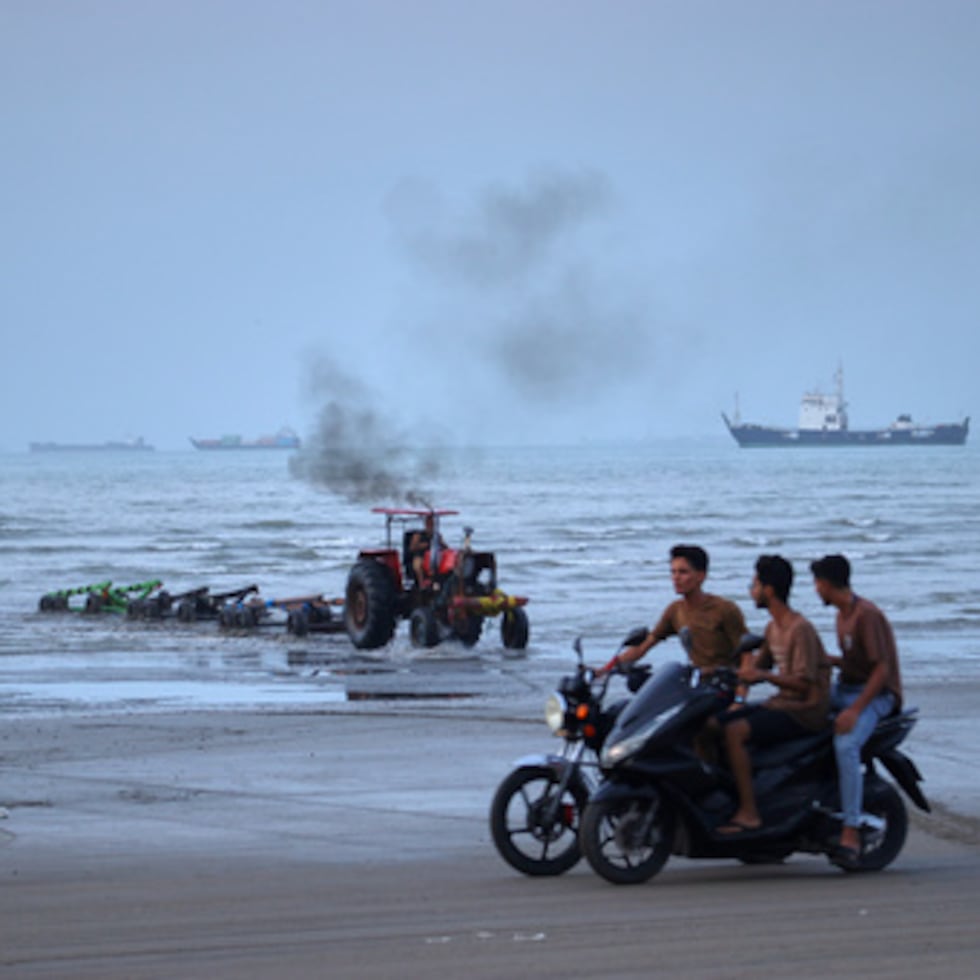 Unos jóvenes circulan en moto por la costa mientras un tractor saca maquinaria del agua y se ven embarcaciones comerciales fondeadas en el estrecho de Ormuz.