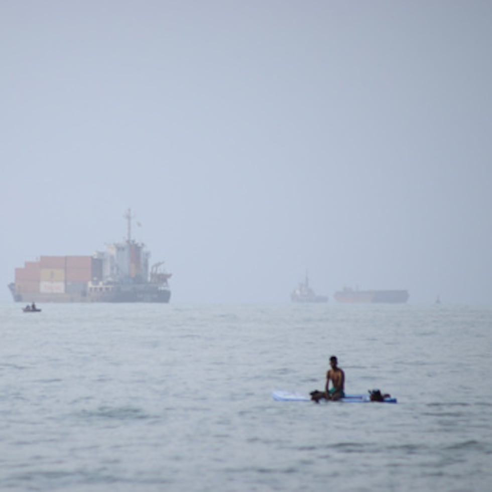 Un hombre está sentado sobre una tabla de surf mientras un buque portacontenedores y otras embarcaciones comerciales aparecen fondeados en el estrecho de Ormuz, frente a Bandar Abbas, en Irán.
