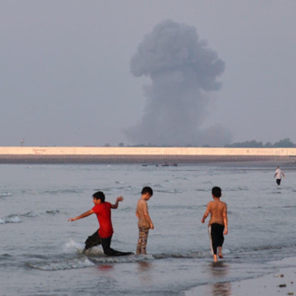 Tres niños juegan en las aguas poco profundas del estrecho de Ormuz, mientras una columna de humo se eleva tras una explosión al fondo.