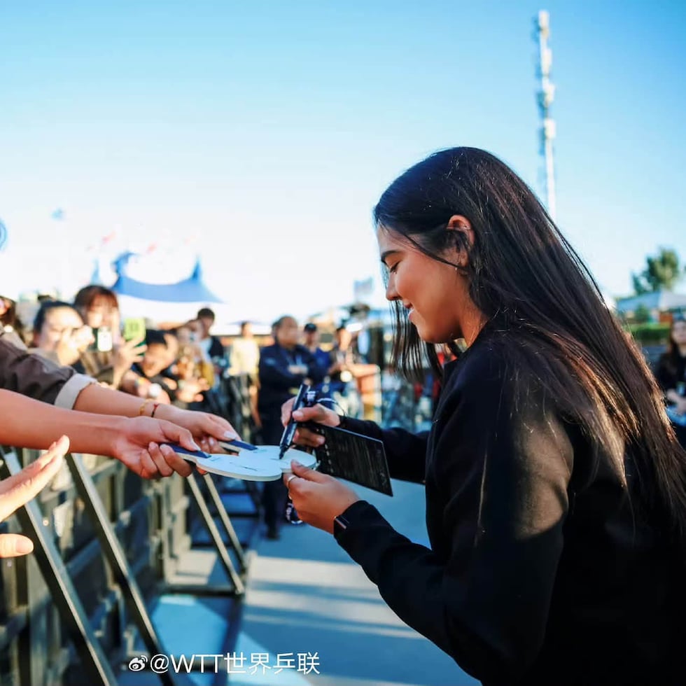 Adriana Díaz appears on a stage signing table tennis rackets and balls, as well as mingling with those in attendance.