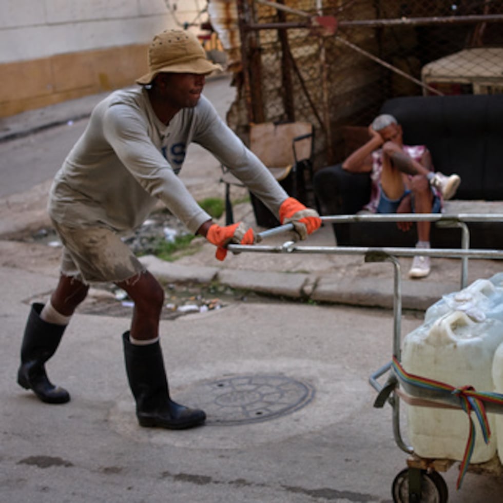 Un hombre empuja un carrito lleno de bidones de agua mientras otro descansa en un sofá en una calle de La Habana, Cuba.