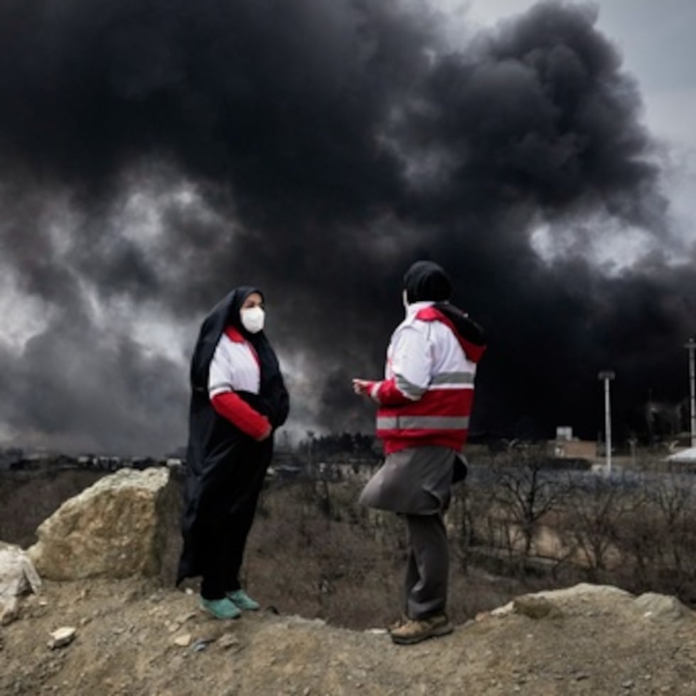 Dos mujeres de la Sociedad de la Media Luna Roja iraní permanecen de pie mientras una densa columna de humo procedente de un ataque estadounidense-israelí contra una instalación de almacenamiento de petróleo.