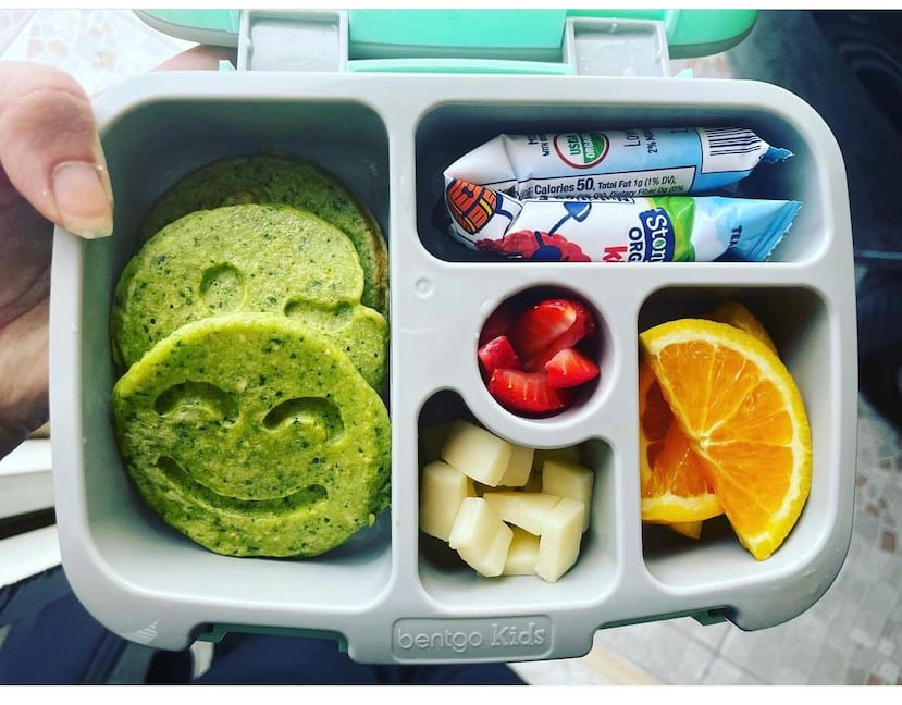 Ejemplo de una lonchera con alimentos saludables que incluye yogurt líquido, pancakes de espinaca, avena y guineo con forma de carita feliz, así como trozos de manzana, china y fresas.