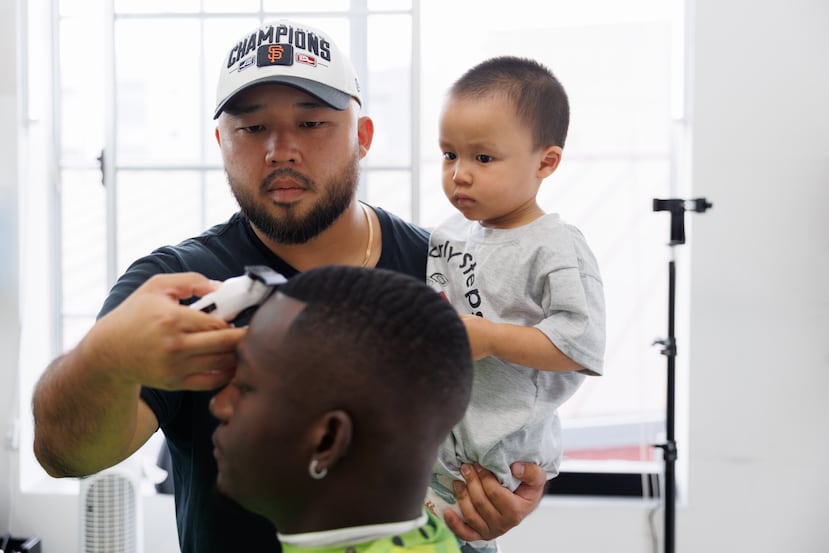 Jack Chang le corta el pelo a Isaac Obamehinti mientras sostiene a su hijo Cairo Miu, de 2 años, en su estudio el lunes 13 de julio de 2026 en San Francisco. (Foto AP/Juliana Yamada)