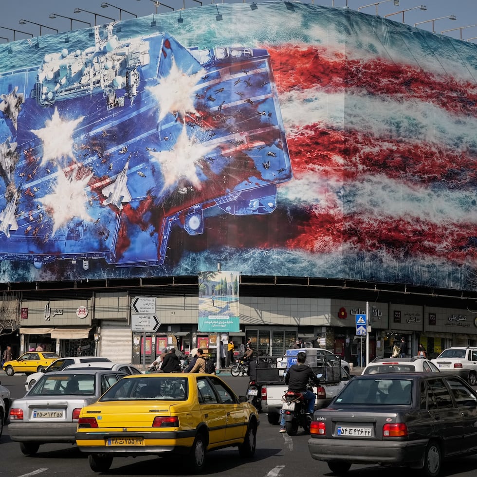 Vehículos pasan junto a una valla publicitaria que representa un portaaviones estadounidense con aviones de combate dañados en la cubierta, y un cartel en farsi e inglés que dice: "Si siembras vientos, cosecharás tempestades", en la plaza Enqelab-e-Eslami (Revolución Islámica) el domingo, 22 de febrero de 2026, en Teherán, Irán. (AP Foto/Vahid Salemi)