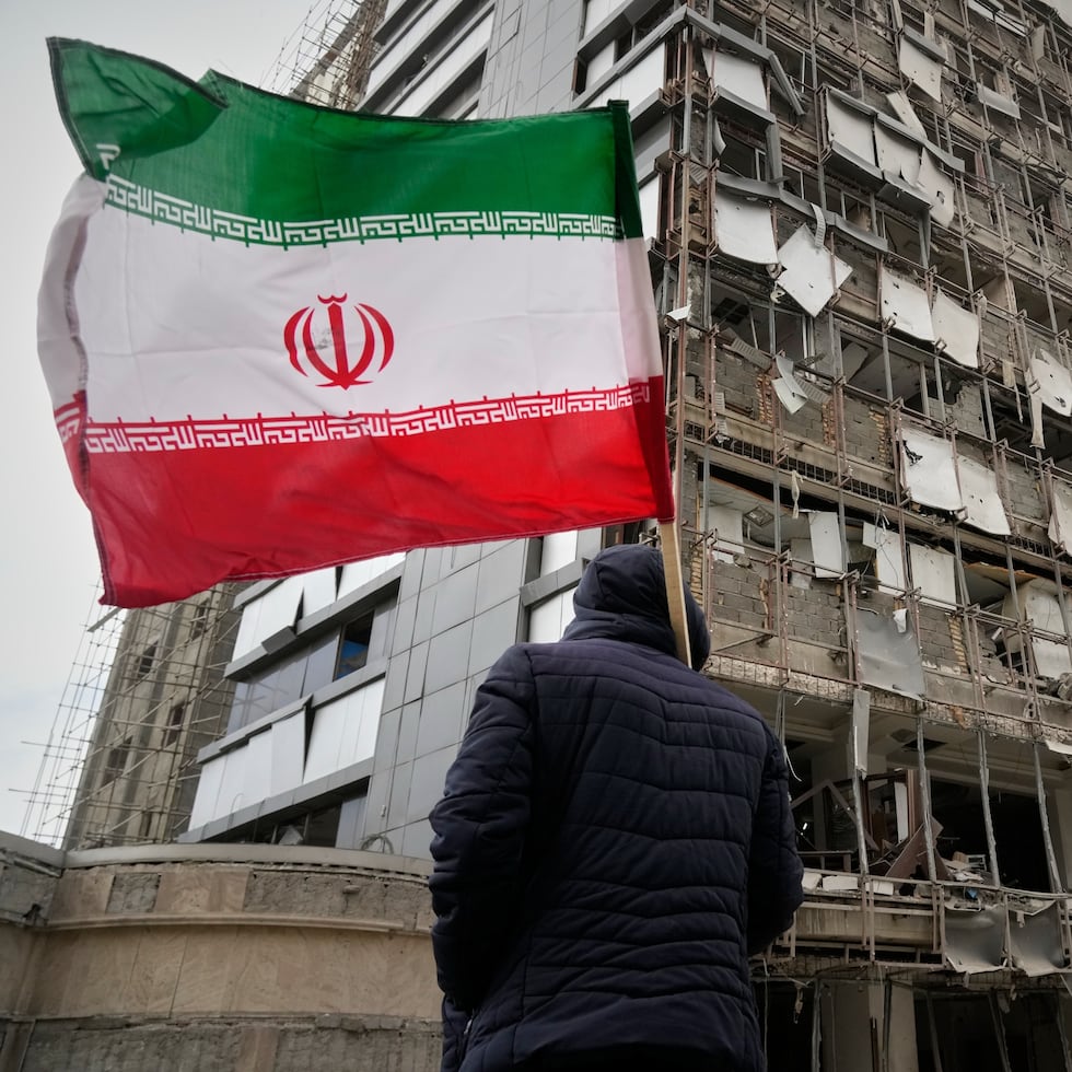 Un hombre sostiene una bandera iraní mientras mira la fachada dañada de Hospital Gandhi, alcanzado el domingo por un ataque que también golpeó una torre de comunicaciones de la televisora estatal y edificios cercanos al otro lado de la calle, durante una campaña de bombardeos estadounidenses e israelíes, en Teherán.