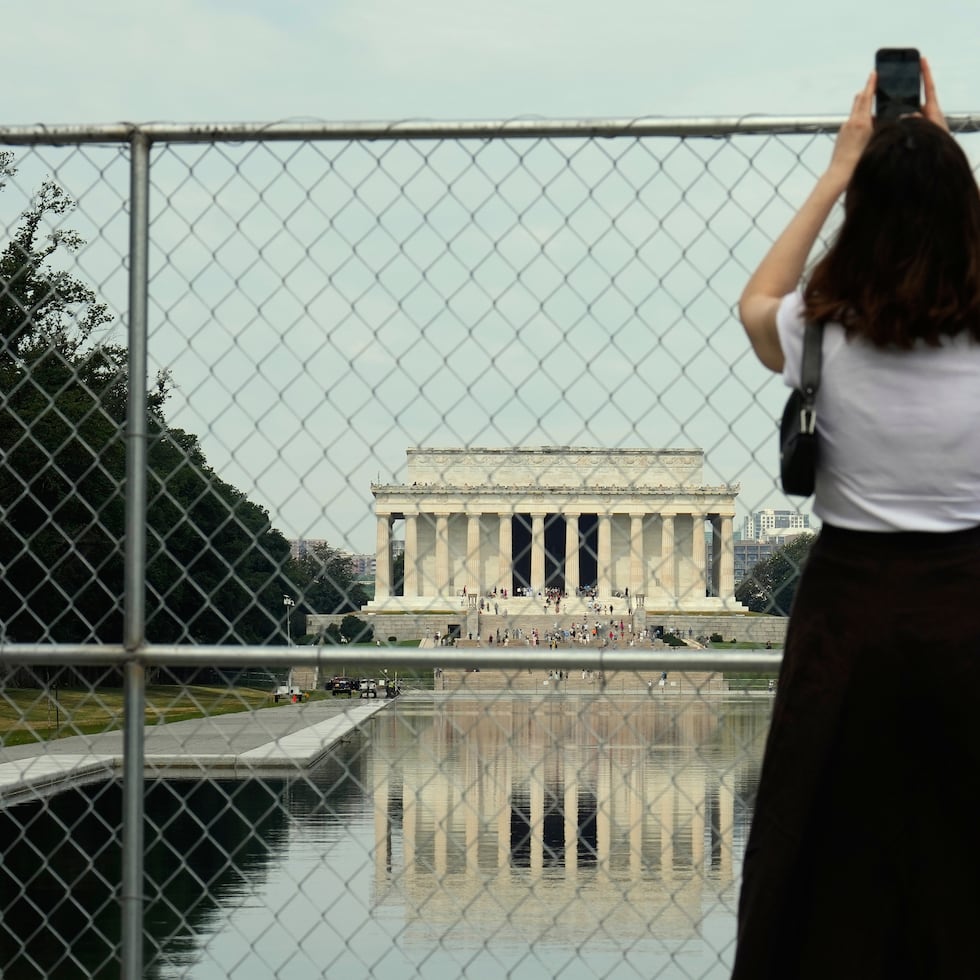 Una persona sostiene un teléfono celular en alto para tomar una foto del estanque reflectante del Monumento a Lincoln y del propio monumento desde detrás de una valla de tela metálica.