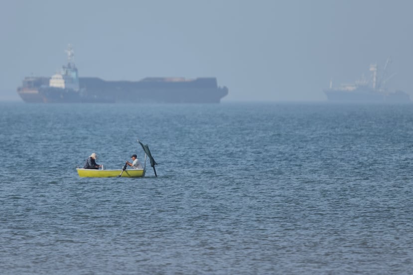 Dos hombres se sientan en una pequeña embarcación sobre el agua mientras buques de carga permanecen anclados al fondo en el estrecho de Ormuz, frente a Bandar Abbas, Irán, el lunes 4 de mayo de 2026. (Amirhosein Khorgooi/ISNA vía AP)