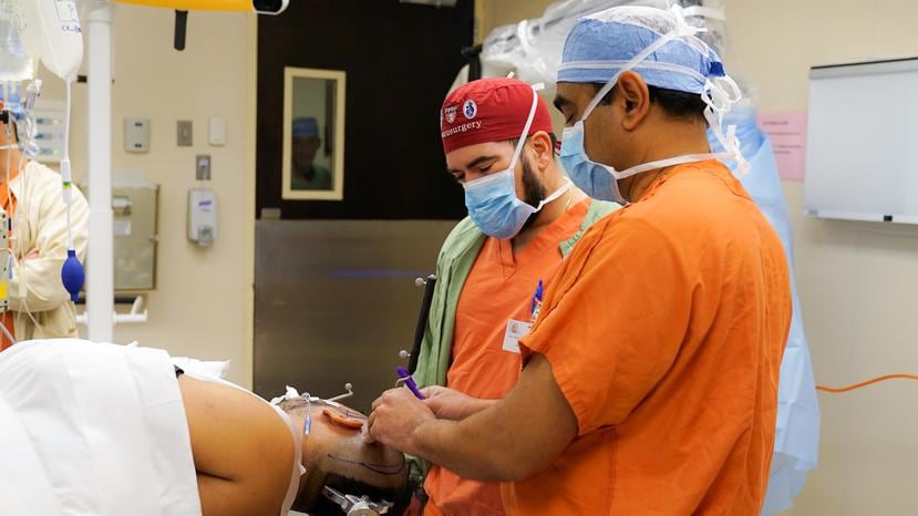El doctor Nirav Patel traza con su marcador quirúrgico, en la cabeza del paciente sedado, la ruta que debe seguir el cirujano Rodolfo Alcedo Guardia al cortar la piel.