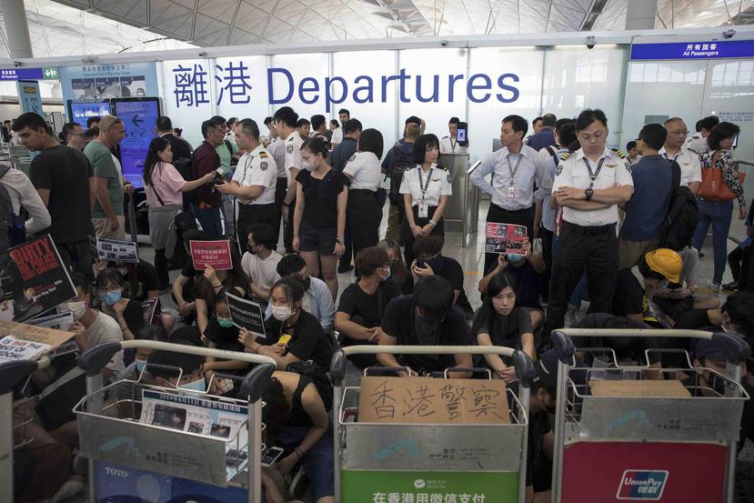 Personal de seguridad del aeropuerto de Hong Kong hace guardia mientras viajeros pasan junto a manifestantes durante una sentada en la terminal de salidas, en Hong Kong. (AP)